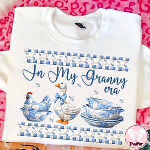 May include: White sweatshirt with a blue and white patchwork design. The text "In My Granny era" is written in blue script. The design includes a chicken, a goose with a blue bow, and a serving dish.