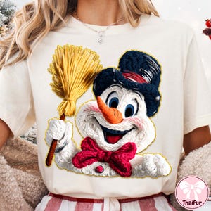 May include: Cream t-shirt with a smiling snowman graphic. The snowman has a carrot nose, blue eyes, a black hat, and a red bow tie. He holds a yellow broom. The graphic is outlined in gold.