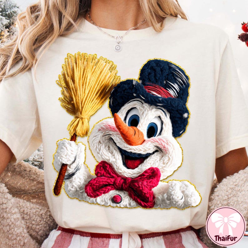 May include: Cream t-shirt with a smiling snowman graphic. The snowman has a carrot nose, blue eyes, a black hat, and a red bow tie. He holds a yellow broom. The graphic is outlined in gold.