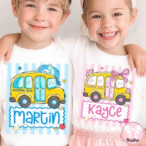 May include: Two white t-shirts with school bus graphics. One shirt has the name "Martin" in blue, the other has "Kayce" in pink. Both shirts feature a yellow school bus design with blue and pink accents, stars, and flowers.
