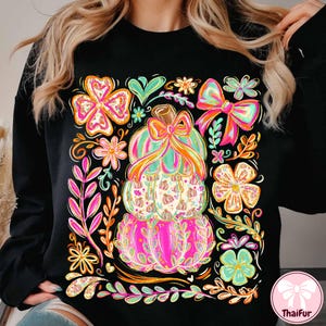 May include: Black sweatshirt featuring a stacked pumpkin design with a colorful bow and floral accents. The pumpkins are pink, green, and orange, with a decorative border of flowers and leaves. The design is vibrant and eye-catching.