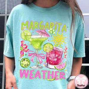 May include: A light blue t-shirt with a margarita-themed graphic. The design features two cocktails, lime slices, a pink bow, and the words "Margarita Weather" in green and pink lettering. A sun and flowers are also included.