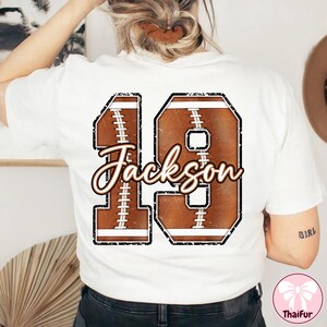 May include: White t-shirt featuring a football-themed design with the number 19 and the name "Jackson" in a distressed style. The design incorporates brown and white elements, resembling a football.