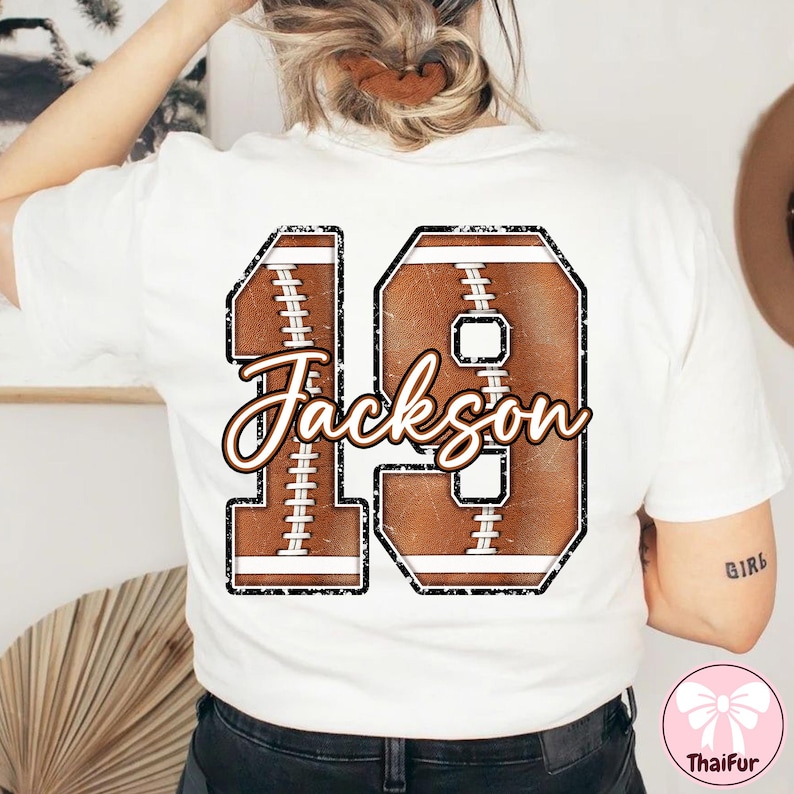 May include: White t-shirt featuring a football-themed design with the number 19 and the name "Jackson" in a distressed style. The design incorporates brown and white elements, resembling a football.