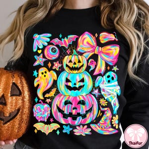 May include: Black sweatshirt featuring a vibrant Halloween-themed design. The graphic includes stacked jack-o'-lanterns, ghosts, bats, bows, and stars in a rainbow of colors. A glittery orange pumpkin is visible on the left.