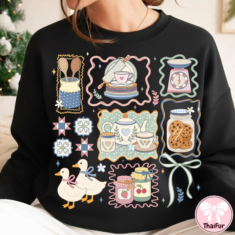 Peut inclure: Sweat-shirt noir avec un motif illustr&eacute; fantaisiste repr&eacute;sentant des &eacute;l&eacute;ments de cuisine et de p&acirc;tisserie. Le motif comprend une th&eacute;i&egrave;re, des tasses &agrave; th&eacute;, des biscuits, des pots de confiture, une balance et deux canards.