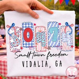 Op de afbeelding: Een wit t-shirt met een afbeelding van het getal 30474 in kleurrijke, gedessineerde blokken met geschulpte randen, versierd met strikken en sterren. Onder de afbeelding staat de tekst Small Town freedom en VIDALIA, GA in zwarte letters.
