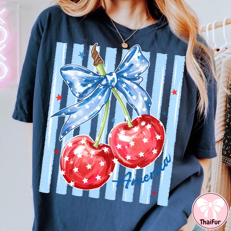 May include: Navy blue t-shirt featuring a patriotic design of two cherries with white stars, a blue bow, and the word "America". The cherries are red, and the bow is blue with white stars. The shirt has a relaxed fit.