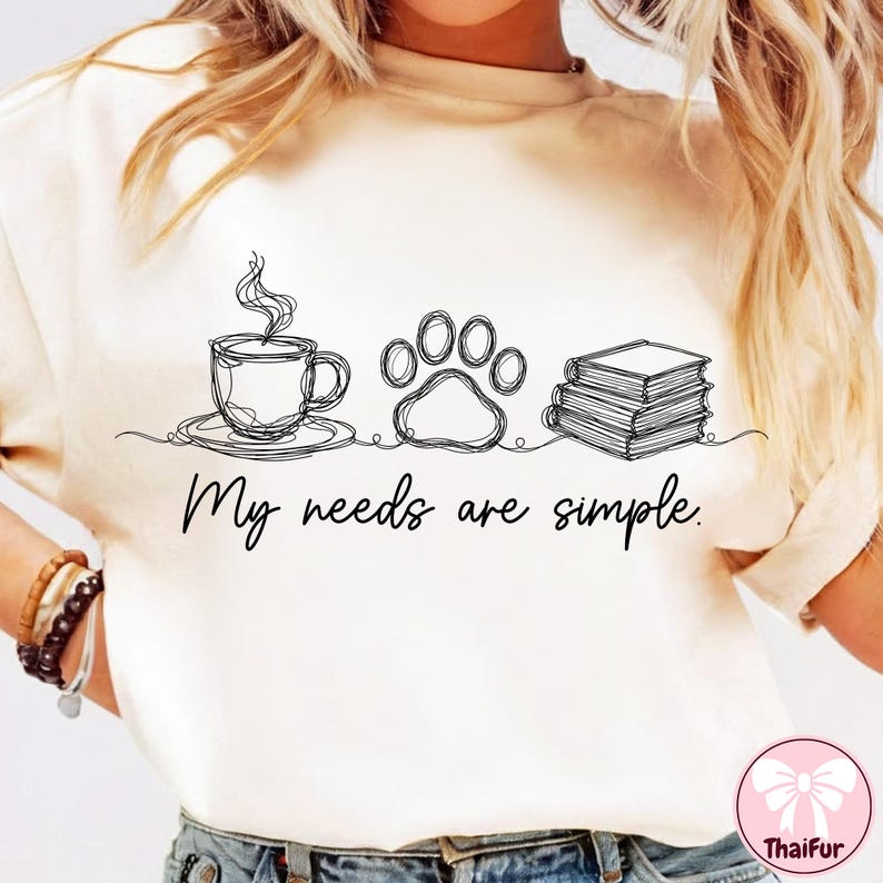 May include: Cream-colored t-shirt with a minimalist line drawing of a coffee cup, a paw print, and a stack of books. The text "My needs are simple" is written below the drawings. The shirt is a casual style.