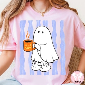 May include: A light pink t-shirt featuring a graphic of a white ghost wearing white slippers and holding an orange jack-o'-lantern mug. The background of the graphic consists of vertical light blue stripes with a distressed, weathered texture.