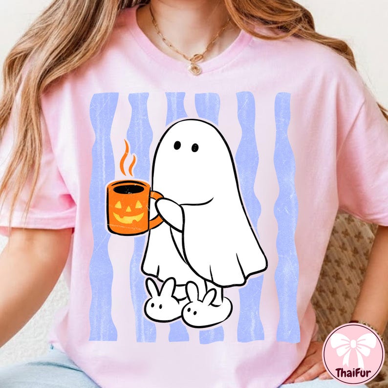 May include: A light pink t-shirt featuring a graphic of a white ghost wearing white slippers and holding an orange jack-o'-lantern mug. The background of the graphic consists of vertical light blue stripes with a distressed, weathered texture.