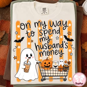 May include: A cream-colored t-shirt featuring orange vertical stripes and the text "on my way to spend my husband's money." The graphic includes a ghost holding a boba tea, a shopping cart with a pumpkin, a candle, and a smaller ghost.