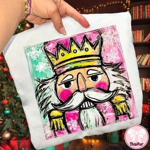 May include: White cushion featuring a colourful illustration of a nutcracker king. The design includes a crown, moustache, and snowflakes on a pink and green background. The cushion is held by a person.