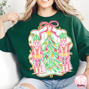 May include: A dark green sweatshirt featuring a festive design. The design includes two nutcrackers, a Christmas tree, and a decorative frame with a bow. The artwork is in shades of pink, green, and gold, creating a holiday theme.