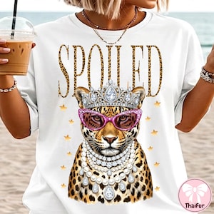 May include: White t-shirt with the word "SPOILED" in leopard print lettering. The shirt features a leopard illustration wearing a crown, pink sunglasses, and a pearl necklace. Gold stars are scattered around the leopard.