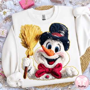 May include: White sweatshirt with a smiling snowman design. The snowman has a carrot nose, blue eyes, and a red bow tie. He is holding a yellow broom. The design is outlined in gold.