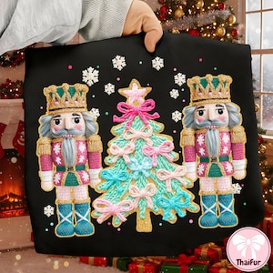 May include: Black pillow cover featuring two nutcrackers and a Christmas tree with pink and blue bows. The nutcrackers wear gold crowns and pink jackets. White snowflakes are scattered around the design. The pillow is held by a person.