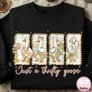 Puede incluir: Sudadera negra con un diseño de cuatro gansos de dibujos animados con delantales de retazos, cada uno enmarcado con un diseño floral. Los gansos están adornados con accesorios y el texto "Just a thrifty goose" está debajo.
