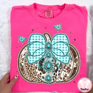 May include: A hot pink Comfort Colors t-shirt featuring a pumpkin design. The pumpkin has a turquoise and white gingham bow, turquoise gemstone accents, and a leopard and cow print design. Three turquoise butterflies are also present.