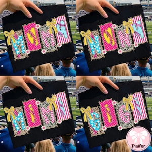 Può includere: T-shirt nero con la scritta "MOM" in lettere colorate e decorate. Le lettere sono decorate con stampa leopardata, stelle, palline da baseball e strisce. Ogni lettera è bordata da un motivo floreale e sormontata da un fiocco giallo.