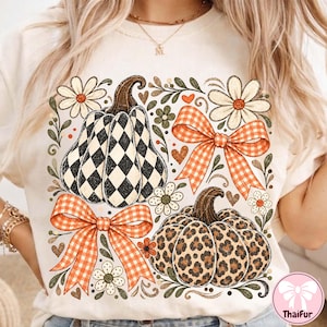 May include: A cream-colored t-shirt featuring a graphic print of two pumpkins, one with a black and white checkered pattern and one with a leopard print. The design includes two orange gingham bows, white daisies, and small green leaves.