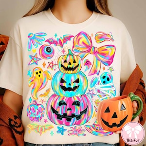 May include: A cream-colored t-shirt featuring a vibrant Halloween design. The design includes stacked jack-o'-lanterns, ghosts, bats, and a large bow, all in a rainbow color scheme. An orange pumpkin mug is held in the hand.