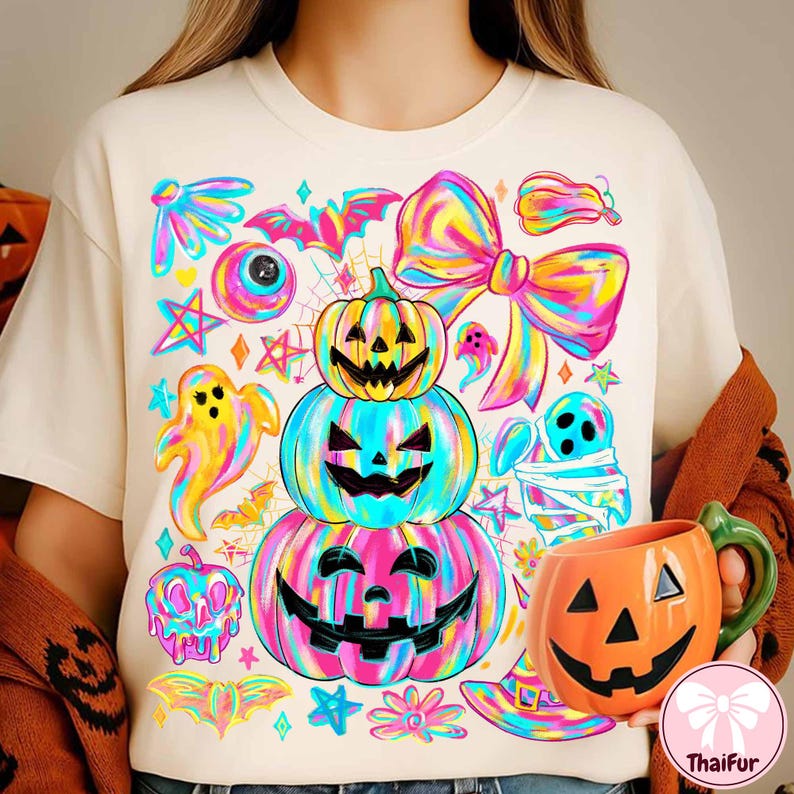 May include: A cream-colored t-shirt featuring a vibrant Halloween design. The design includes stacked jack-o'-lanterns, ghosts, bats, and a large bow, all in a rainbow color scheme. An orange pumpkin mug is held in the hand.