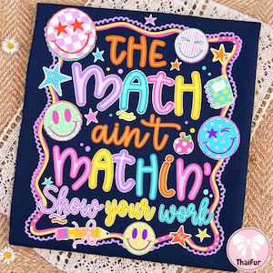 May include: A navy blue t-shirt featuring the colorful text THE MATH ain't MATHIN' Show your work. The design includes various smiley faces, stars, a small notebook, an apple, and a pencil, all surrounded by a wavy pink and yellow border.