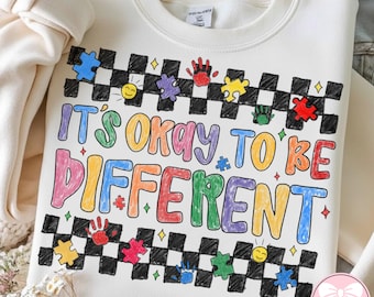 Op de afbeelding: Crèmekleurige sweatshirt met de tekst "IT'S OKAY TO BE DIFFERENT" in kleurrijke, handgeschreven letters. Het ontwerp bevat puzzelstukjes, handafdrukken en een zwart-wit geblokte rand.