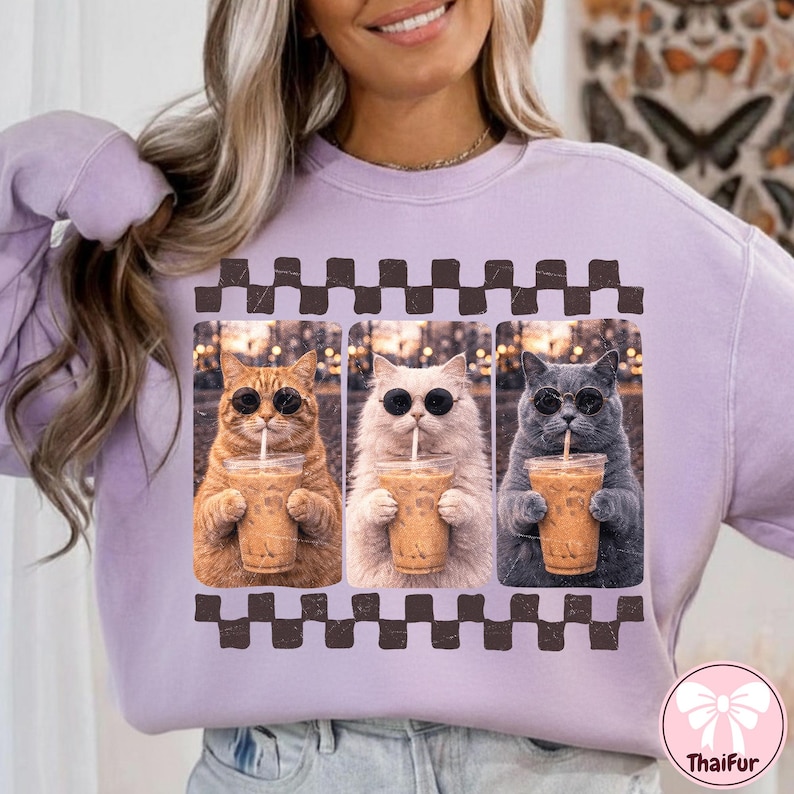 May include: A light purple sweatshirt featuring a graphic of three cats wearing sunglasses and drinking iced coffee. The cats are in a row, each with a different fur color: orange, white, and gray. A brown and white checkered pattern is above the cats.