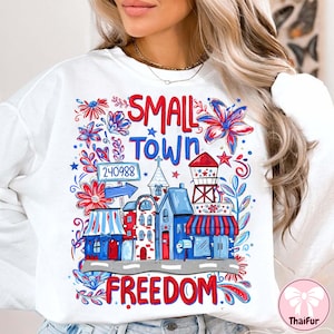 Puede incluir: Sudadera blanca con un diseño gráfico colorido. El diseño presenta las palabras "SMALL TOWN FREEDOM" con una ilustración de un pueblo pequeño en rojo, blanco y azul. Incluye un código postal.