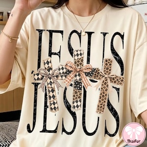 May include: A cream-colored t-shirt featuring the word JESUS printed twice in a large, distressed black font. Between the words are three crosses with frayed edges, each decorated with a different pattern and a small fabric bow at the center.