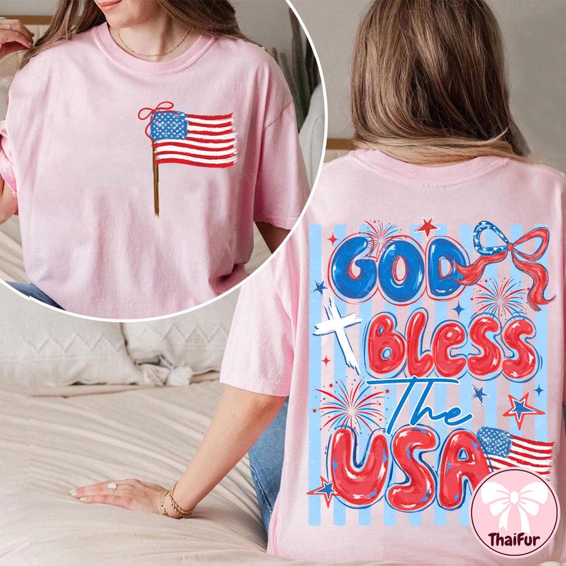 Op de afbeelding: Zachtroze T-shirt met een Amerikaanse vlag op de voorkant en de tekst "God Bless The USA" op de achterkant. Het ontwerp gebruikt rood, wit en blauw, met vuurwerk en sterren.