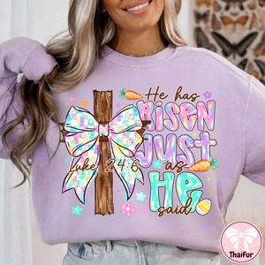 May include: A light purple sweatshirt with a colorful graphic featuring a wooden cross, a bow, carrots, flowers, and the text "He has Risen JUST as HE said" and "Luke 24:6".