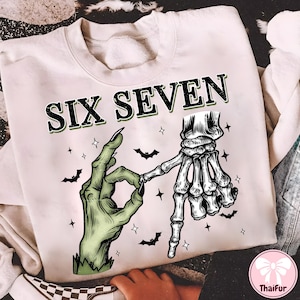 May include: Cream-colored sweatshirt with the text "SIX SEVEN" in black and green. The graphic features a green zombie hand making an "okay" sign, connected to a skeleton hand. Small black bats and stars are scattered around the design.