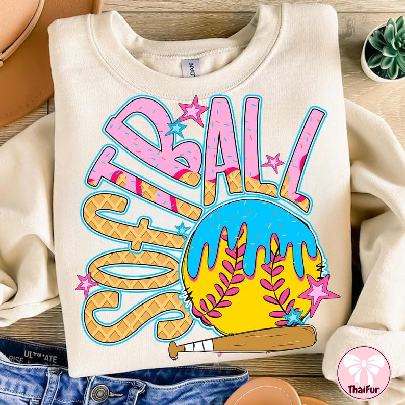 May include: A cream colored sweatshirt with a graphic design of a softball with the word "Softball" written around it. The softball is yellow with pink and blue dripping icing and a green leaf design. The word "Softball" is written in a waffle cone font with pink and blue sprinkles.