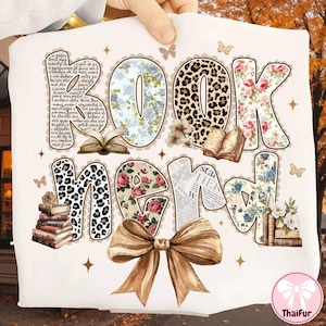 May include: A white graphic tee with the words "BOOK NERD" in a variety of decorative fonts. The letters feature floral, leopard print, and newspaper patterns. A large brown bow is at the bottom, with books and butterflies scattered around.