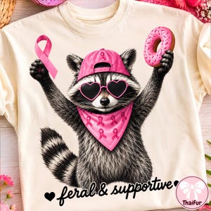 Pode incluir: Uma t-shirt cor de creme com o gráfico de um guaxinim usando um guaxinim a usar um boné de basebol rosa, óculos de sol em forma de coração e uma bandana rosa com padrões de fitas. O guaxinim segura uma fita rosa e um donut rosa. Texto: feral & supportive.