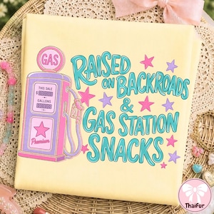 K&ouml;nnte beinhalten: Ein hellgelbes T-Shirt mit einer Grafik einer vintage Zapfs&auml;ule in Lila und Pink. Der Text "RAISED ON BACKROADS & GAS STATION SNACKS" ist in t&uuml;rkisfarbener Schrift gedruckt, umgeben von kleinen rosa und violetten Sternen auf dem Stoff.