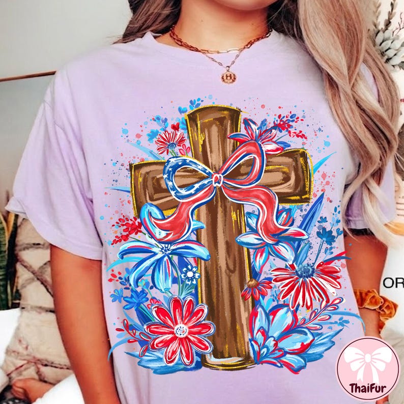 May include: A light purple t-shirt featuring a brown cross adorned with a red, white, and blue bow. The cross is surrounded by red, white, and blue floral accents, creating a patriotic and artistic design.