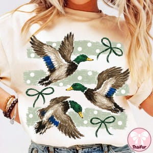 May include: A cream-colored t-shirt featuring a graphic print of three flying mallard ducks in shades of brown, green, and blue. The design includes horizontal sage green brushstroke stripes with white polka dots and dark green ribbon bows.