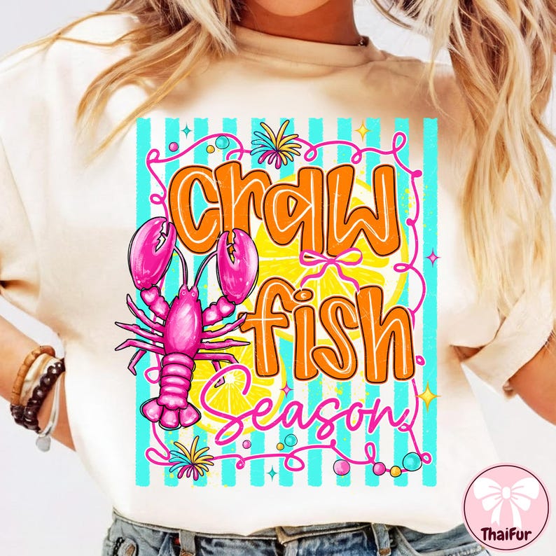 May include: Cream-colored t-shirt with a colorful graphic. The graphic features the words "Crawfish Season" in orange and pink, along with a pink crawfish and lemon slices. The design is set against a backdrop of blue and white stripes.