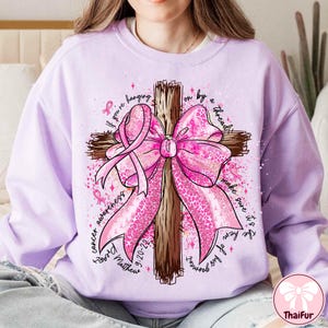 May include: A light purple sweatshirt featuring a wooden cross with a large pink bow and pink ribbons. The words "cancer awareness" and "If you're hanging on by a thread, make sure it's the hem of his garment" are written around the cross. The text "9-20-22" and "Matthew" are also visible.