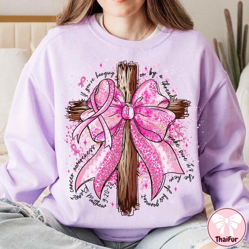 May include: A light purple sweatshirt featuring a wooden cross with a large pink bow and pink ribbons. The words "cancer awareness" and "If you're hanging on by a thread, make sure it's the hem of his garment" are written around the cross. The text "9-20-22" and "Matthew" are also visible.