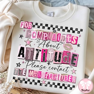 Pode incluir: Camisola branca com o texto "FOR COMPLAINTS ABOUT ATTITUDE Please contact THE MANUFACTURER" em letras rosa, preto e com estampa de leopardo. O design inclui estrelas e uma borda quadriculada.