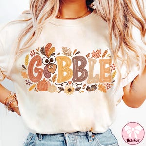 May include: A cream-colored t-shirt featuring the text GOBBLE in large, multi-colored, line-art style letters. The design includes a turkey head replacing the letter O, surrounded by autumn-themed graphics like pumpkins, leaves, and acorns.