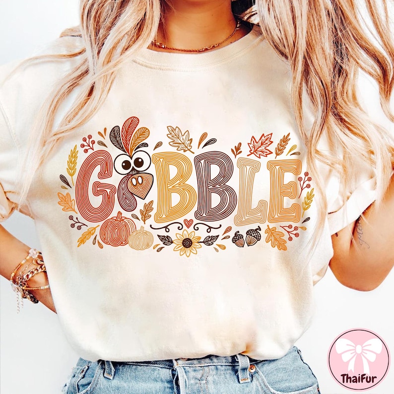May include: A cream-colored t-shirt featuring the text GOBBLE in large, multi-colored, line-art style letters. The design includes a turkey head replacing the letter O, surrounded by autumn-themed graphics like pumpkins, leaves, and acorns.