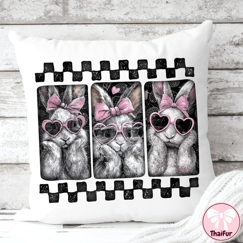 May include: White square pillow featuring three bunny illustrations. Each bunny wears heart-shaped sunglasses and a pink bow. The bunnies are framed by a black and white checkered border. The pillow has a ThaiFur logo in the bottom right corner.