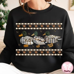 May include: Black sweatshirt featuring three ducks with plaid patterned bodies and green heads. The ducks are arranged in a row, with a decorative border of hearts and plaid above and below them. The sweatshirt has a crew neck.