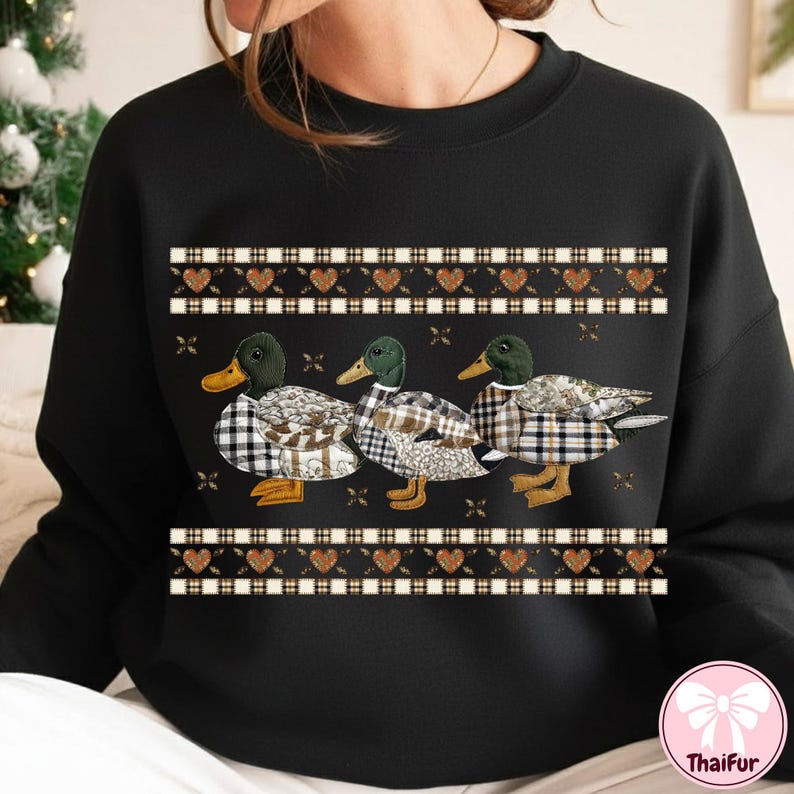 May include: Black sweatshirt featuring three ducks with plaid patterned bodies and green heads. The ducks are arranged in a row, with a decorative border of hearts and plaid above and below them. The sweatshirt has a crew neck.